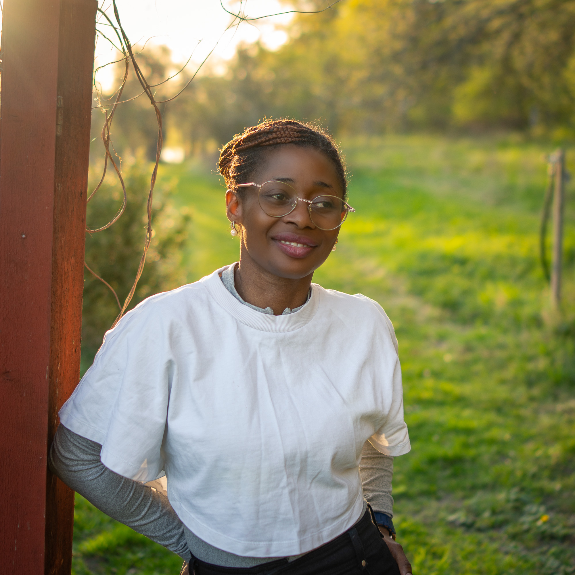 Jamirah smiling in a white tee by a red wooden post in a sunlit Swedish garden