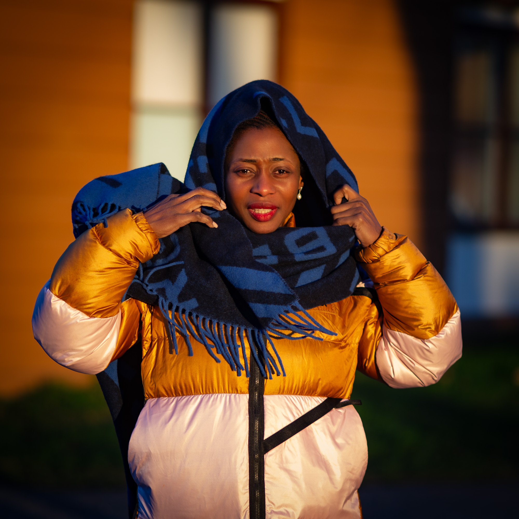 Jamirah in a gold puffer jacket pulling a blue scarf over her head in golden light