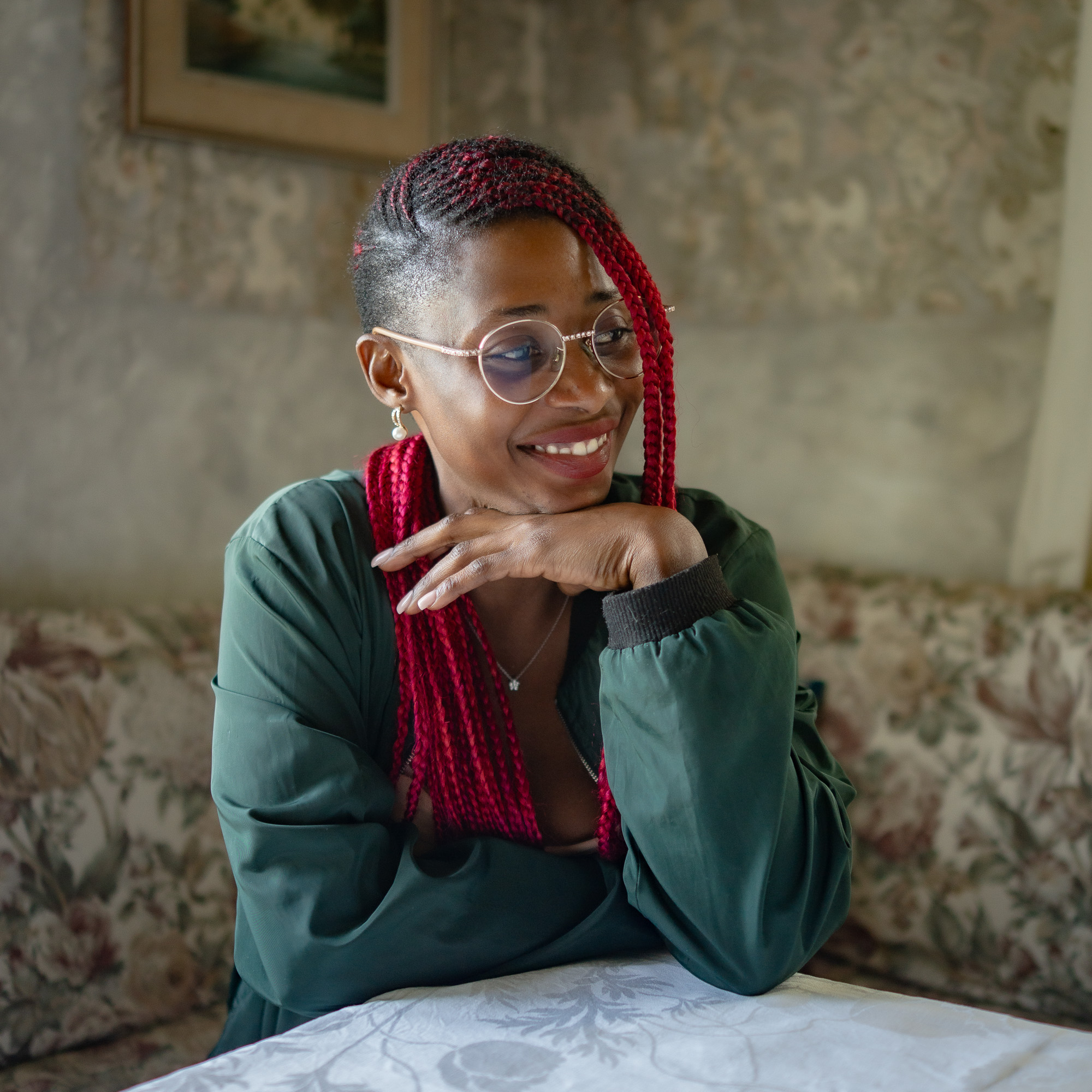 Jamirah smiling indoors in a green shirt with red braids, resting her chin on her hand
