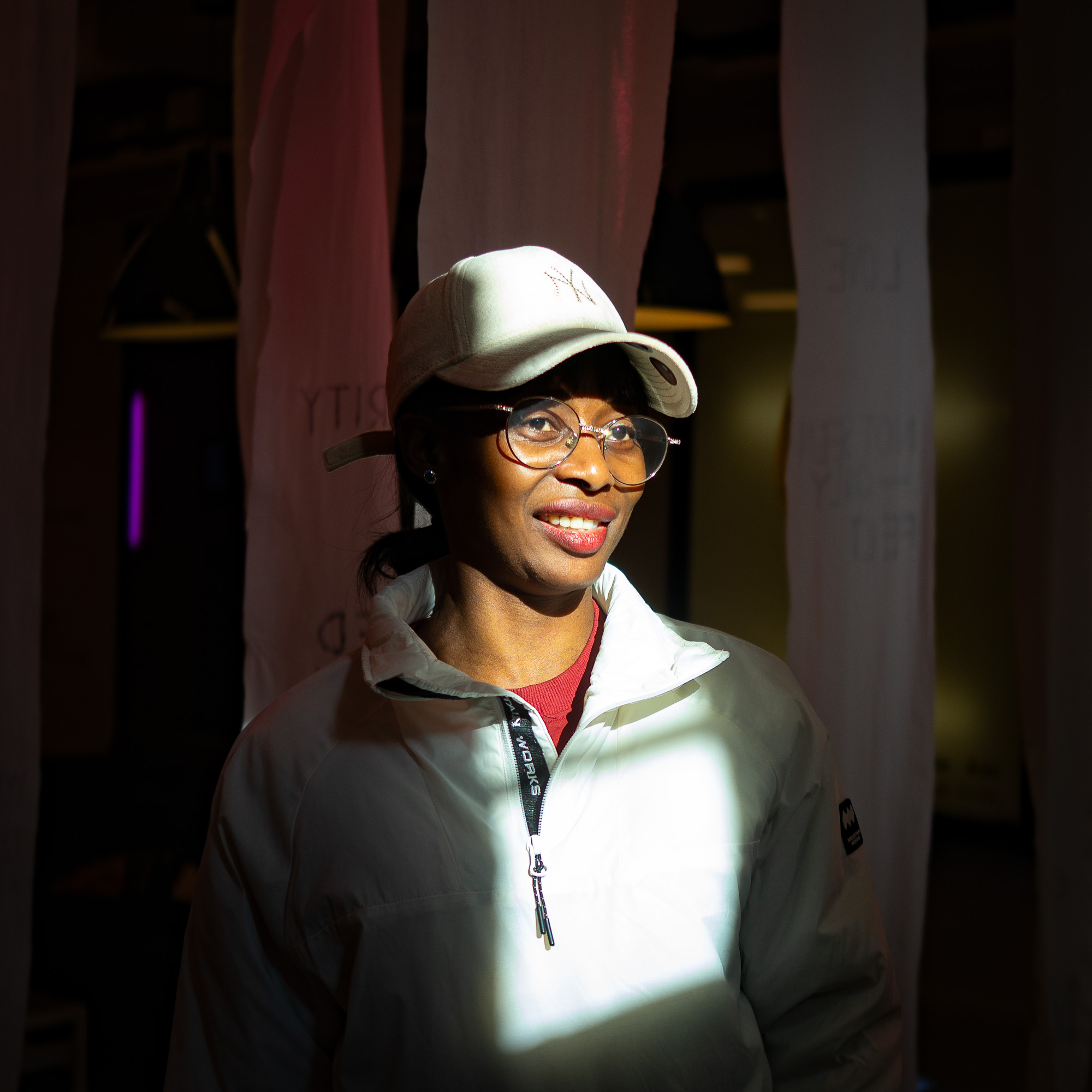 Jamirah smiling in a cream cap and jacket, lit by a beam of light at an evening event